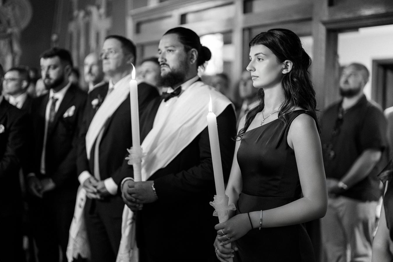 A group of people holding candles during a religious ceremony, dressed in formal attire, with focused expressions, in a church setting.