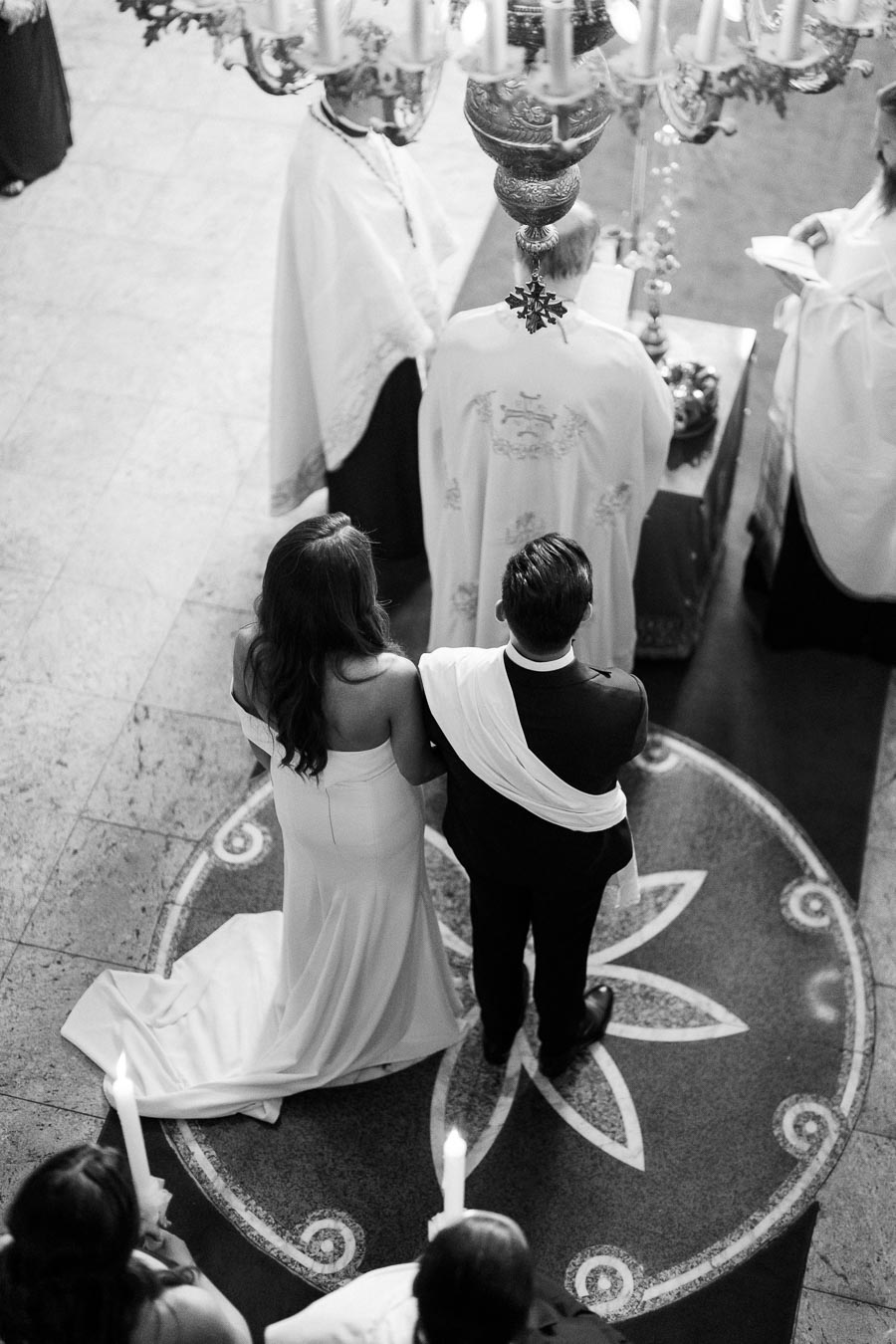 A bride and groom stand side by side at an altar during a traditional wedding ceremony, surrounded by clergy in ornate robes, under an elaborate chandelier, captured in black and white.