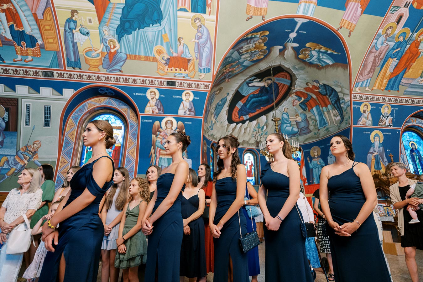 A group of women in matching navy blue dresses stand attentively inside a church adorned with vibrant religious murals and frescoes. The scene is serene and richly decorated, capturing a moment of solemnity and tradition.