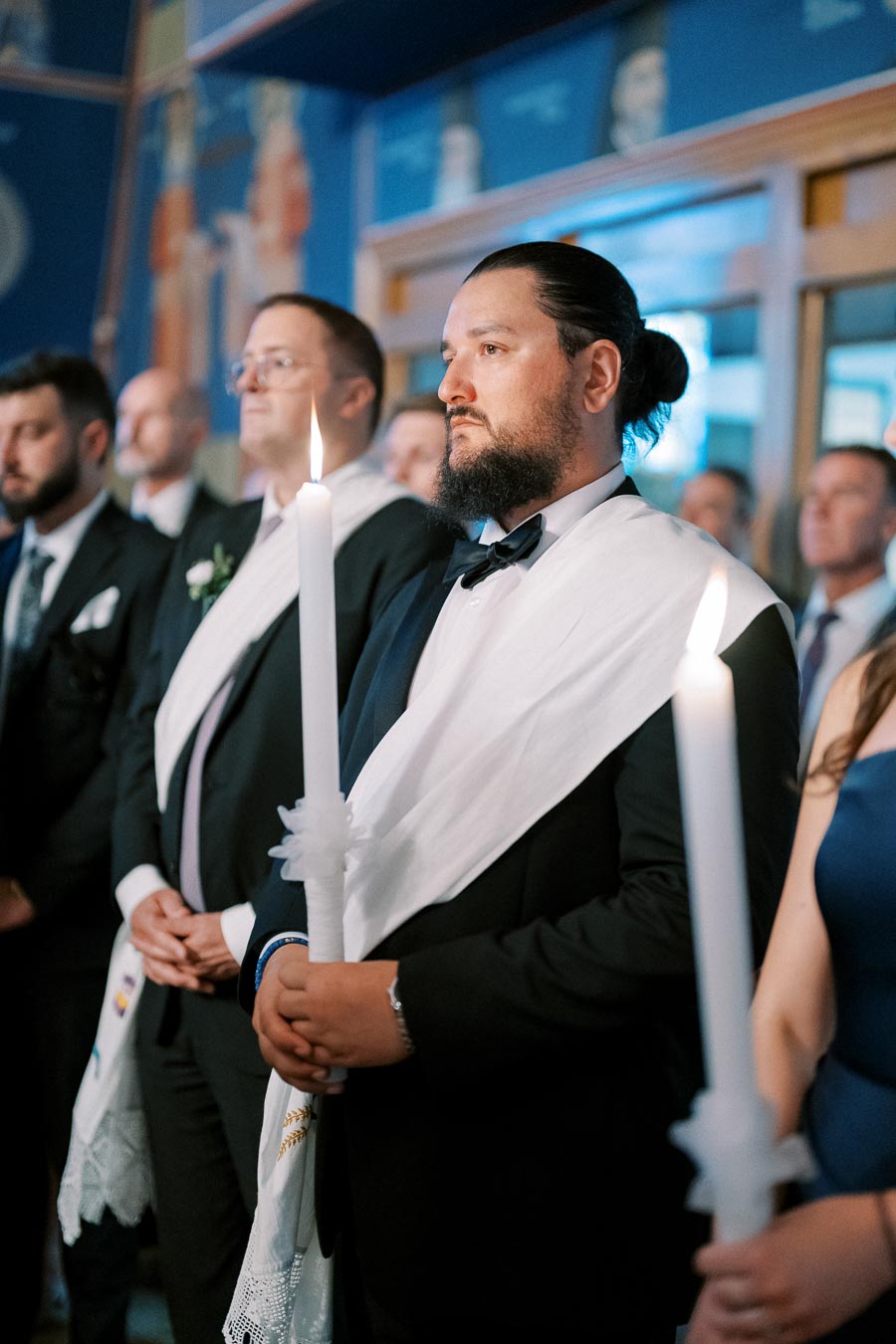 Group of formally dressed individuals holding candles during a ceremonial event, with blue and decorative background.