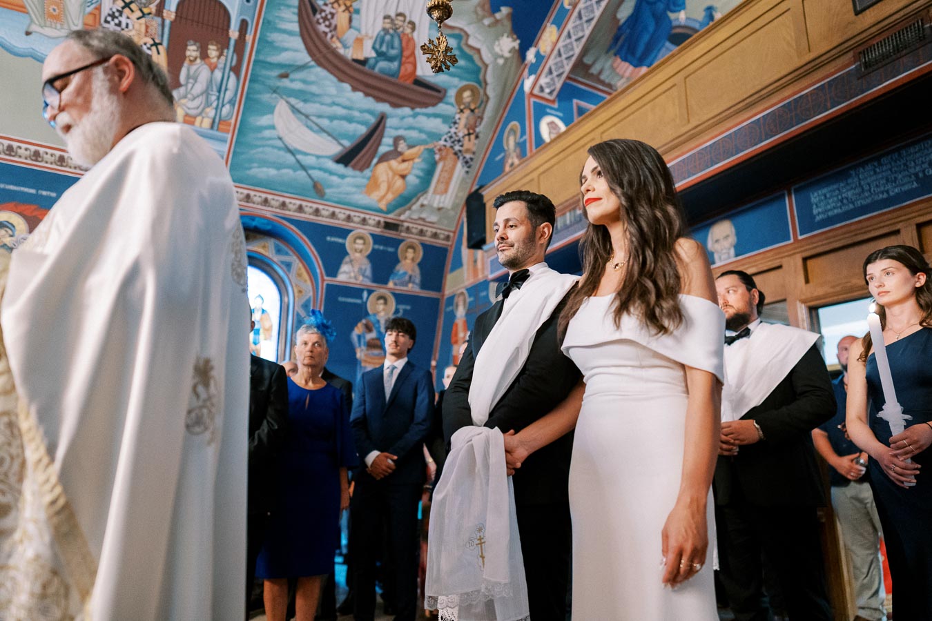 A wedding ceremony inside a richly decorated church, featuring a couple standing solemnly at the altar with a priest in the foreground and guests observing in the background.