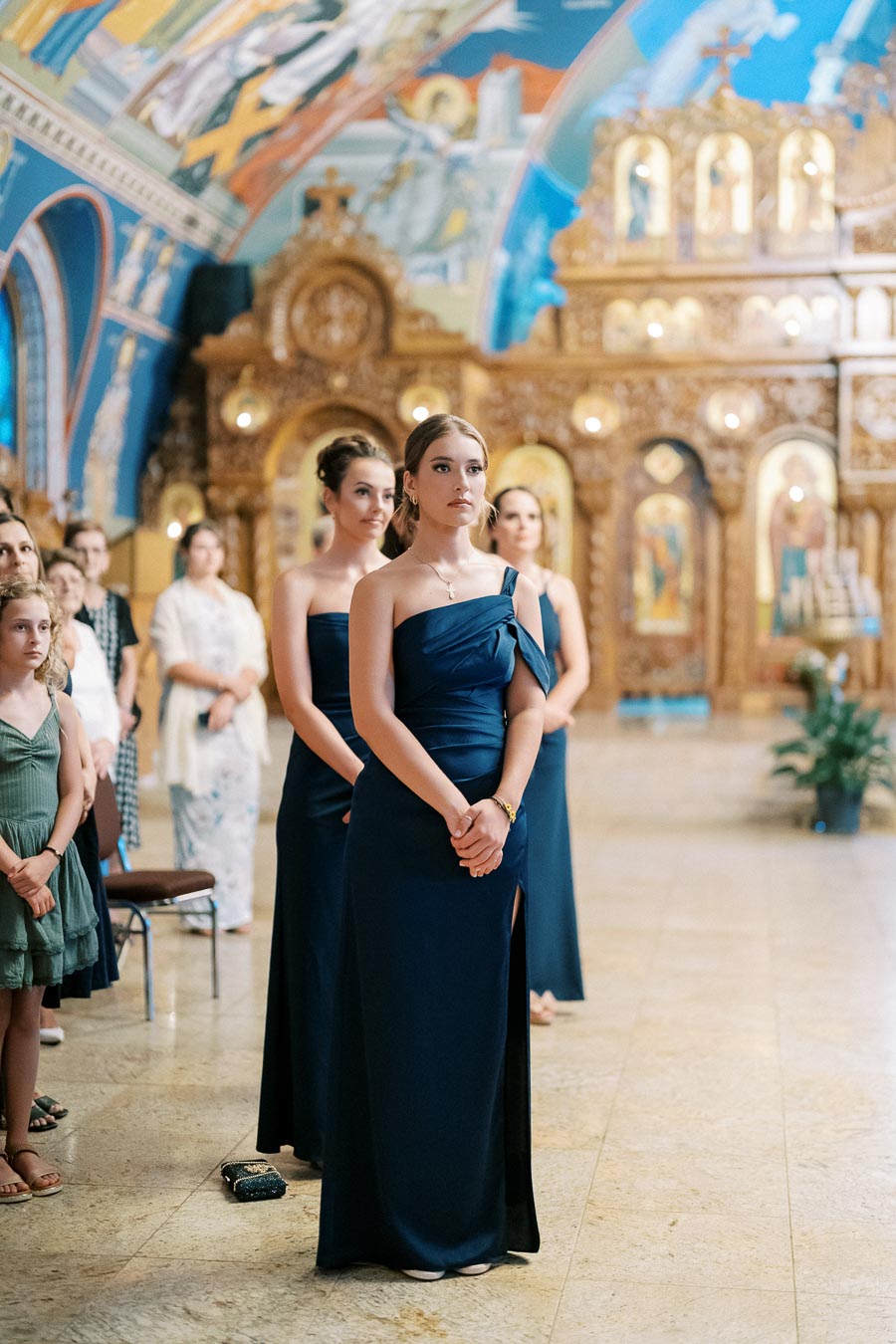 Bridesmaids in elegant navy blue dresses standing inside an ornate church during a wedding ceremony, surrounded by attendees with intricate frescoes and icons in the background.