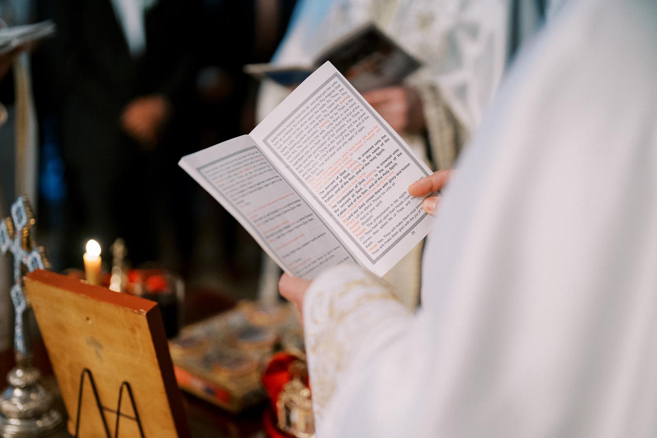 A person in ceremonial attire holding an open prayer booklet during a religious ceremony, with a lit candle and religious artifacts visible in the background.