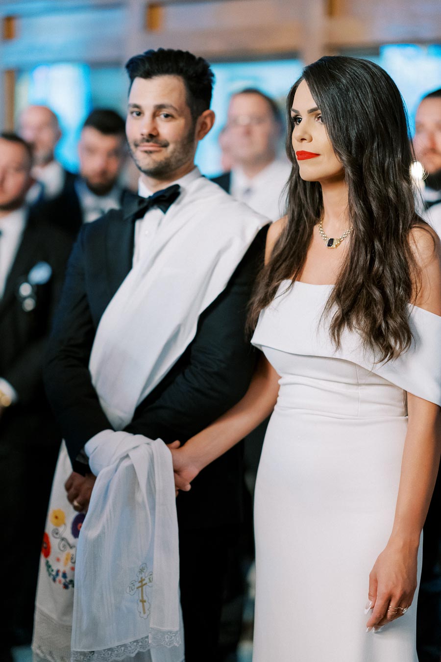 A couple dressed elegantly in formal attire, holding hands during a wedding ceremony, surrounded by guests.