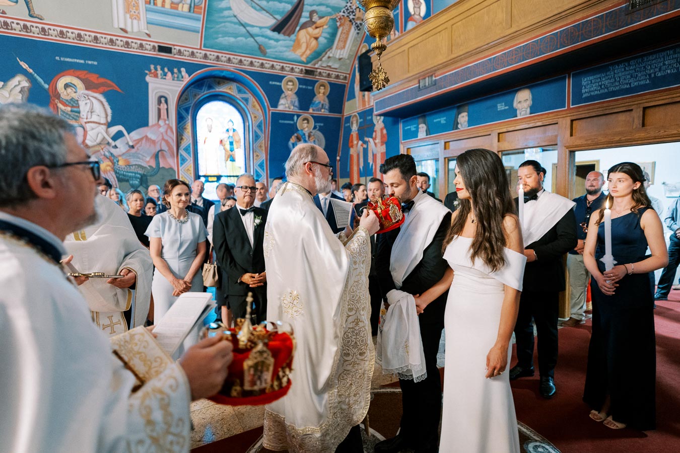 Religious wedding ceremony inside an ornately decorated church, featuring a priest in traditional vestments performing a blessing for a couple holding crowns, surrounded by guests and vivid mural art.