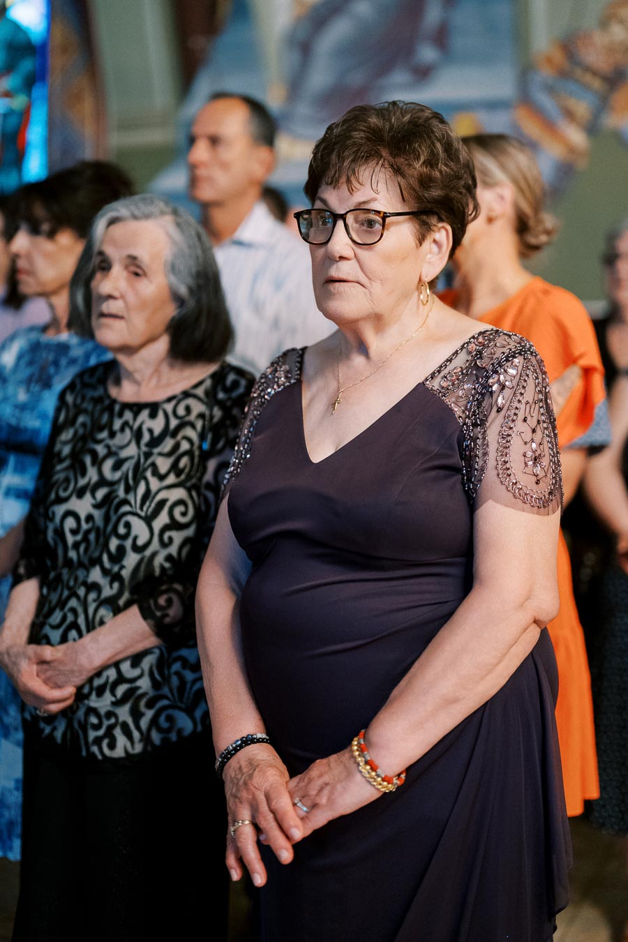 A group of people attentively listening during an indoor event, with two elderly women in focus wearing elegant dresses.