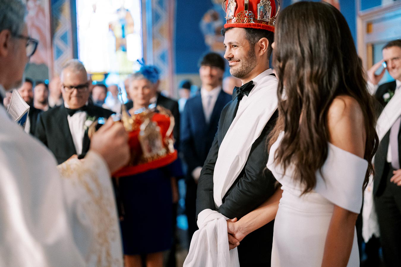 A couple wearing crowns and formal attire participates in a traditional wedding ceremony inside a decorated church, surrounded by guests and a priest.
