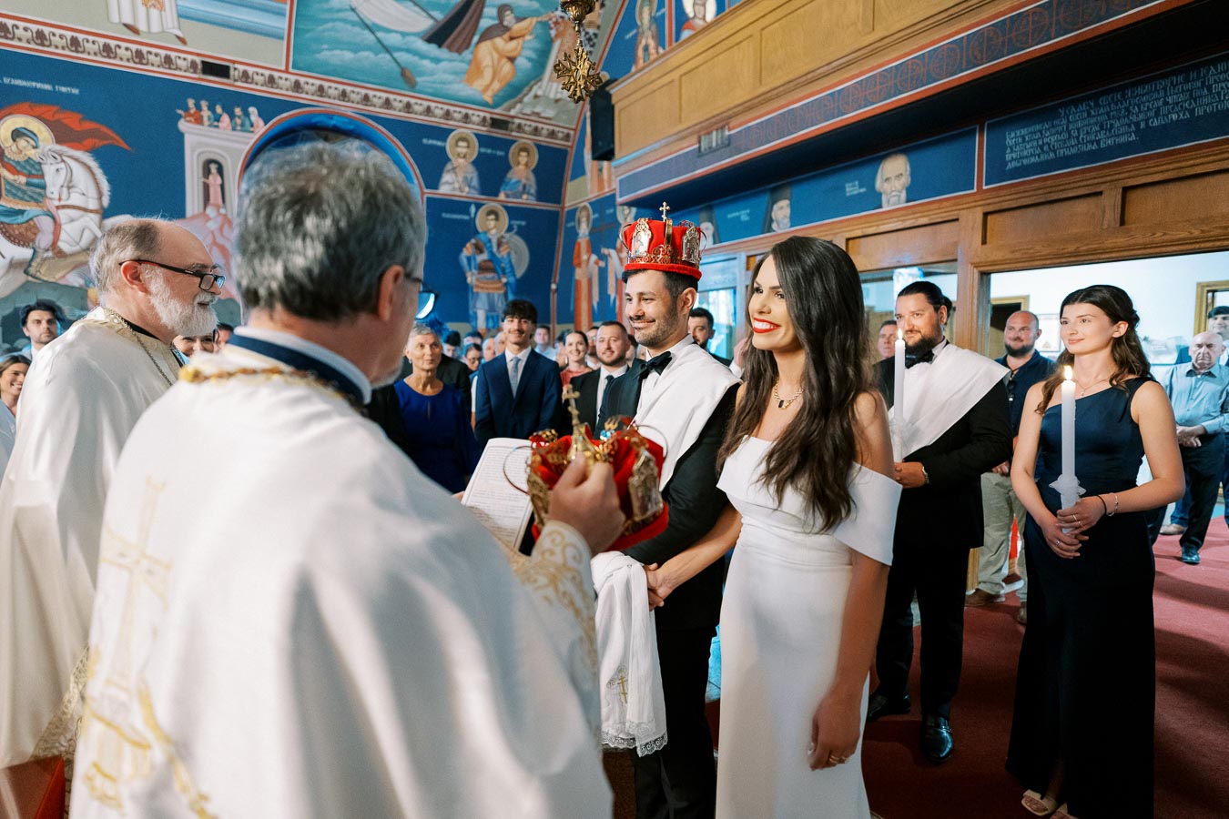 A couple getting married in a traditional religious ceremony inside a beautifully adorned church, surrounded by guests and clergy, with murals and iconography on the walls.