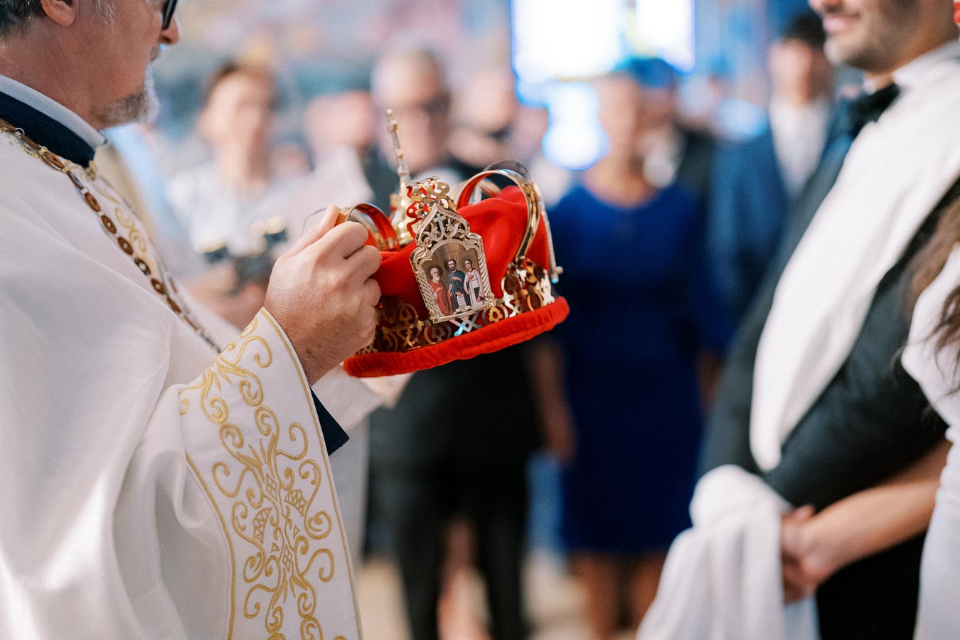 A priest holds an ornate red and gold crown during a traditional wedding ceremony, with blurred guests in the background.