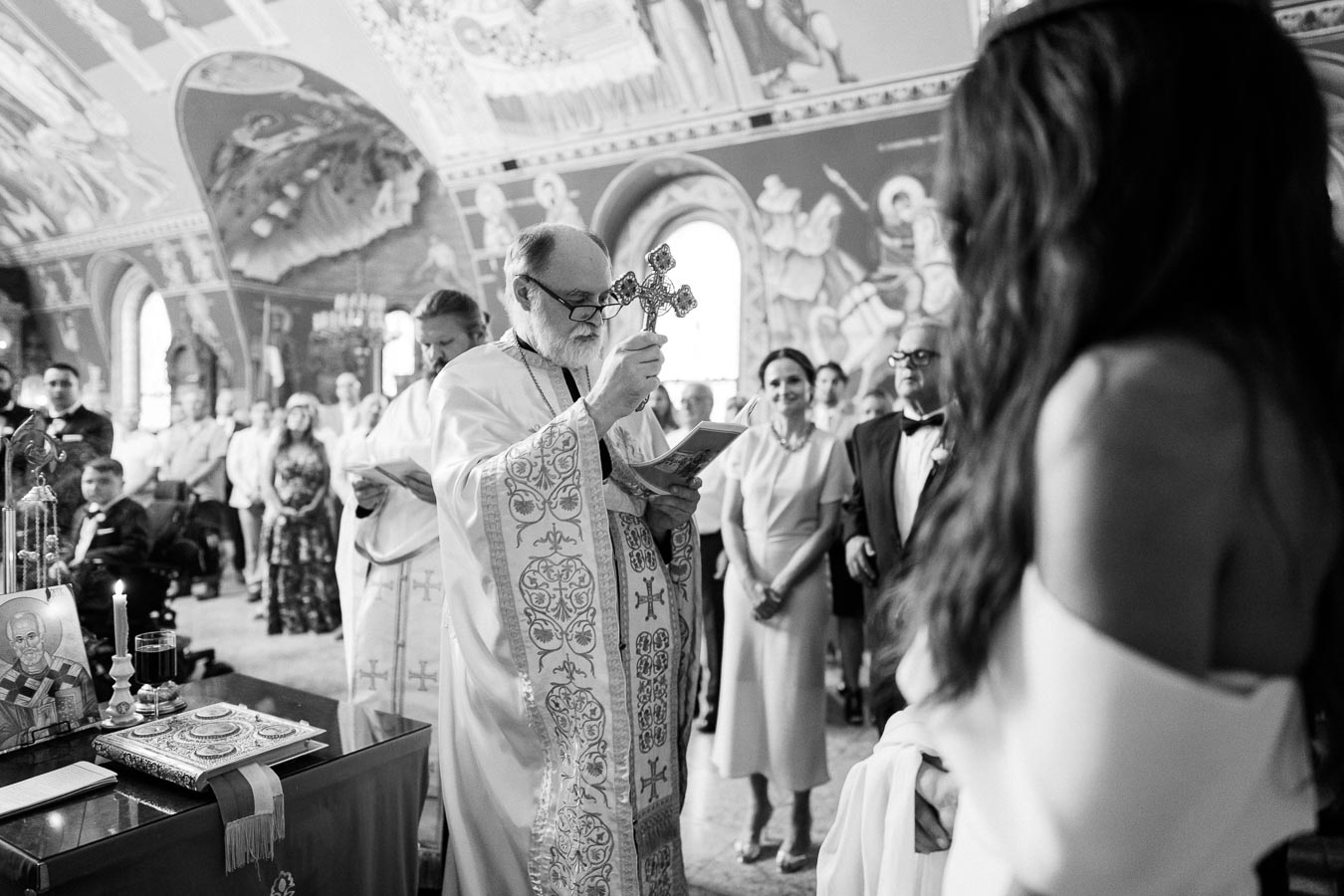 Black and white image of a traditional religious ceremony inside an ornately decorated church, featuring a priest holding a cross while a congregation watches attentively.