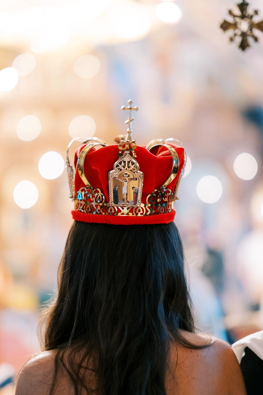 A woman wearing an ornate red crown with a cross at a ceremonial event, with blurred lights in the background.