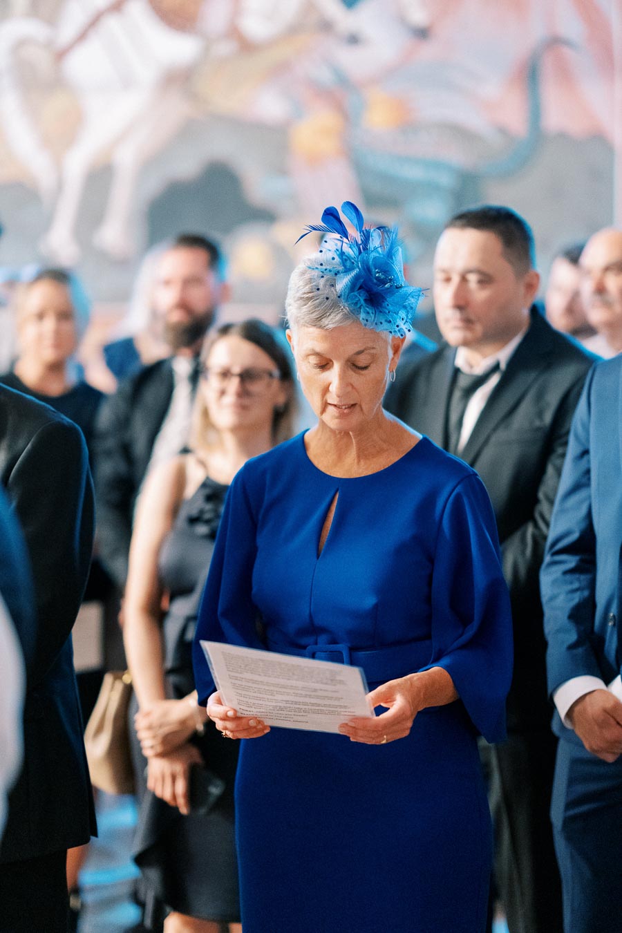 An elegant woman in a vibrant blue dress and matching fascinator reading a speech at a formal event, with a blurred crowd dressed in formal attire in the background.