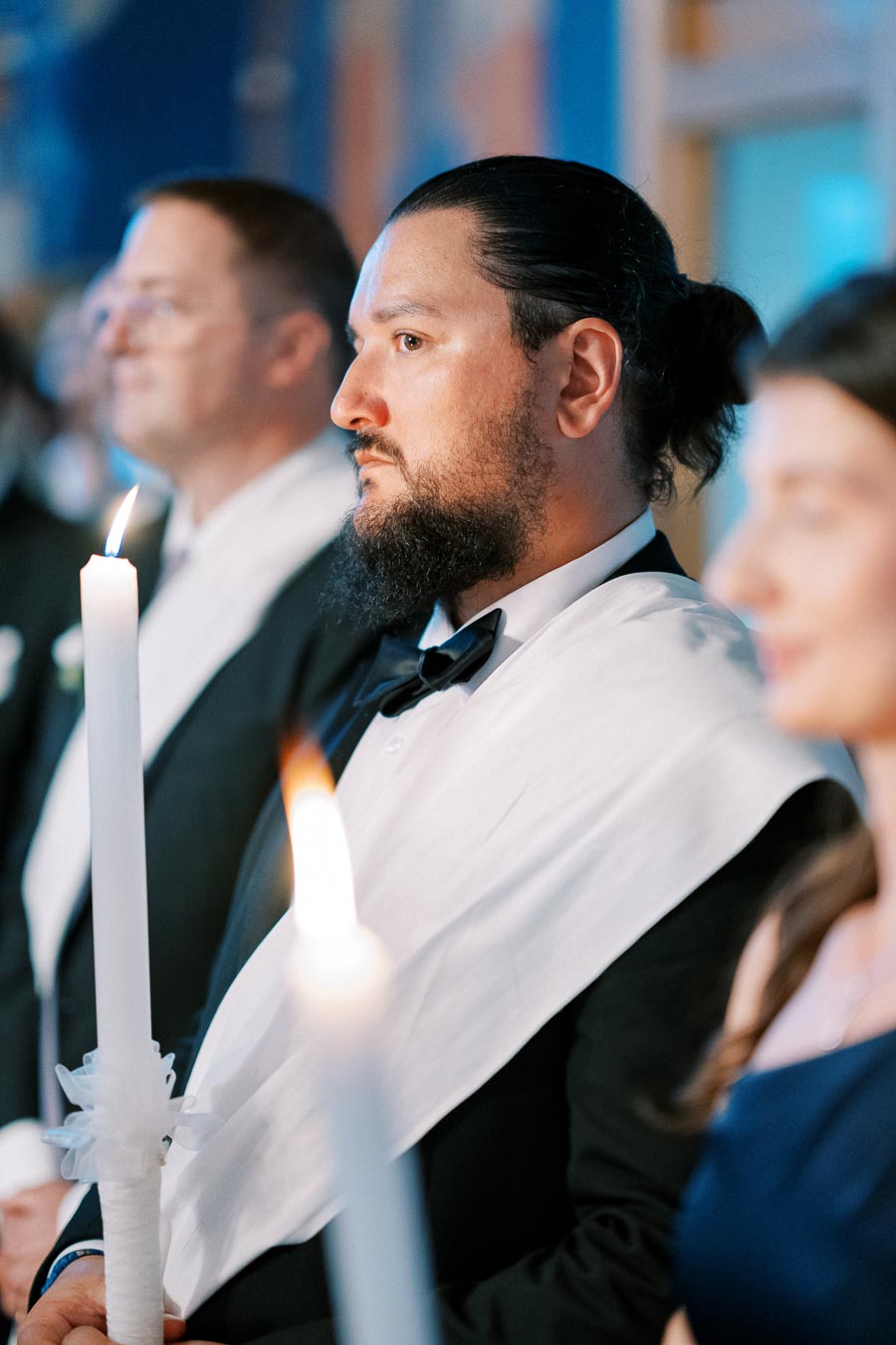 A solemn man in a tuxedo and white shawl holds a lit candle during a formal ceremony, with other participants visible in the background.