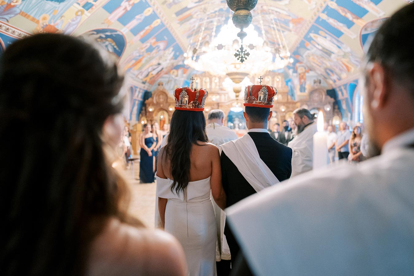 Couple wearing crowns during traditional Orthodox wedding ceremony in ornately decorated church, surrounded by guests and clergy