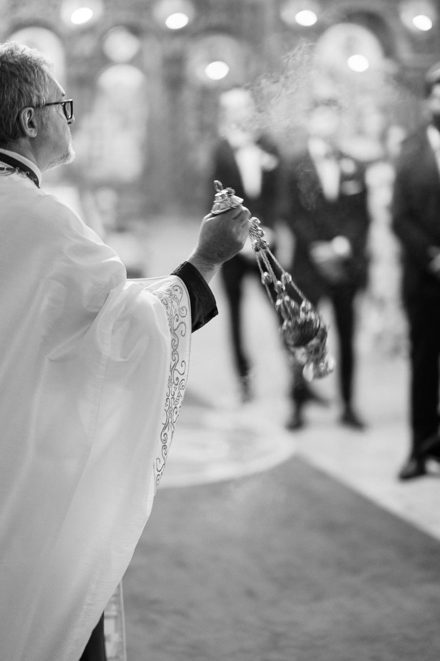A clergy member performs a traditional incense ceremony in a church setting, with blurred figures and ornate decor in the background, emphasizing a spiritual and solemn atmosphere.