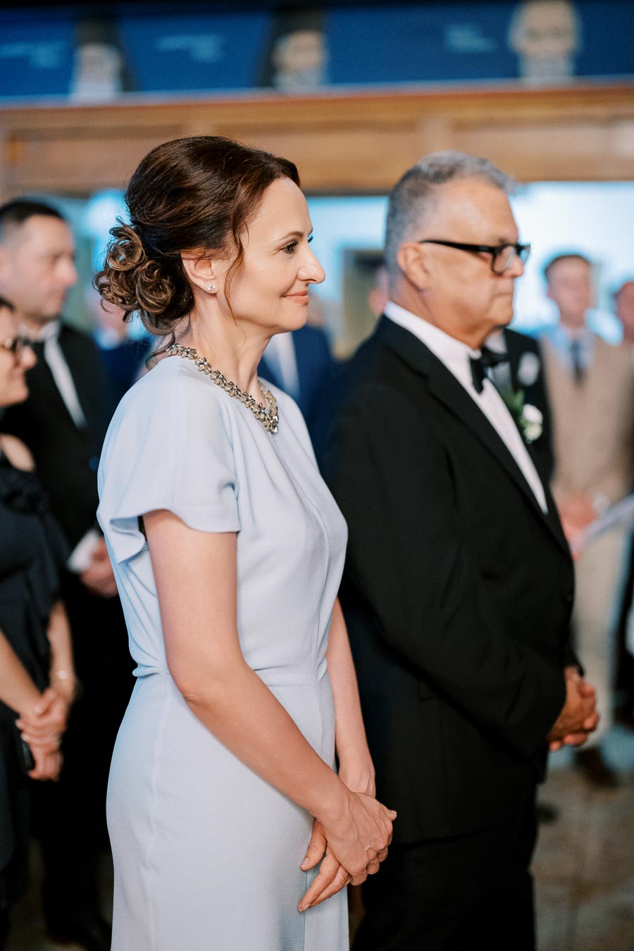Elegant woman and man in formal attire attending a ceremony, surrounded by a group of people in a sophisticated setting.