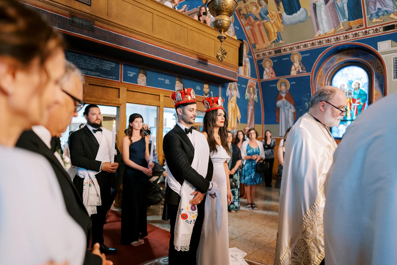 A couple stands side by side wearing crowns during a traditional wedding ceremony in an ornately decorated church, with guests and a priest in attendance.