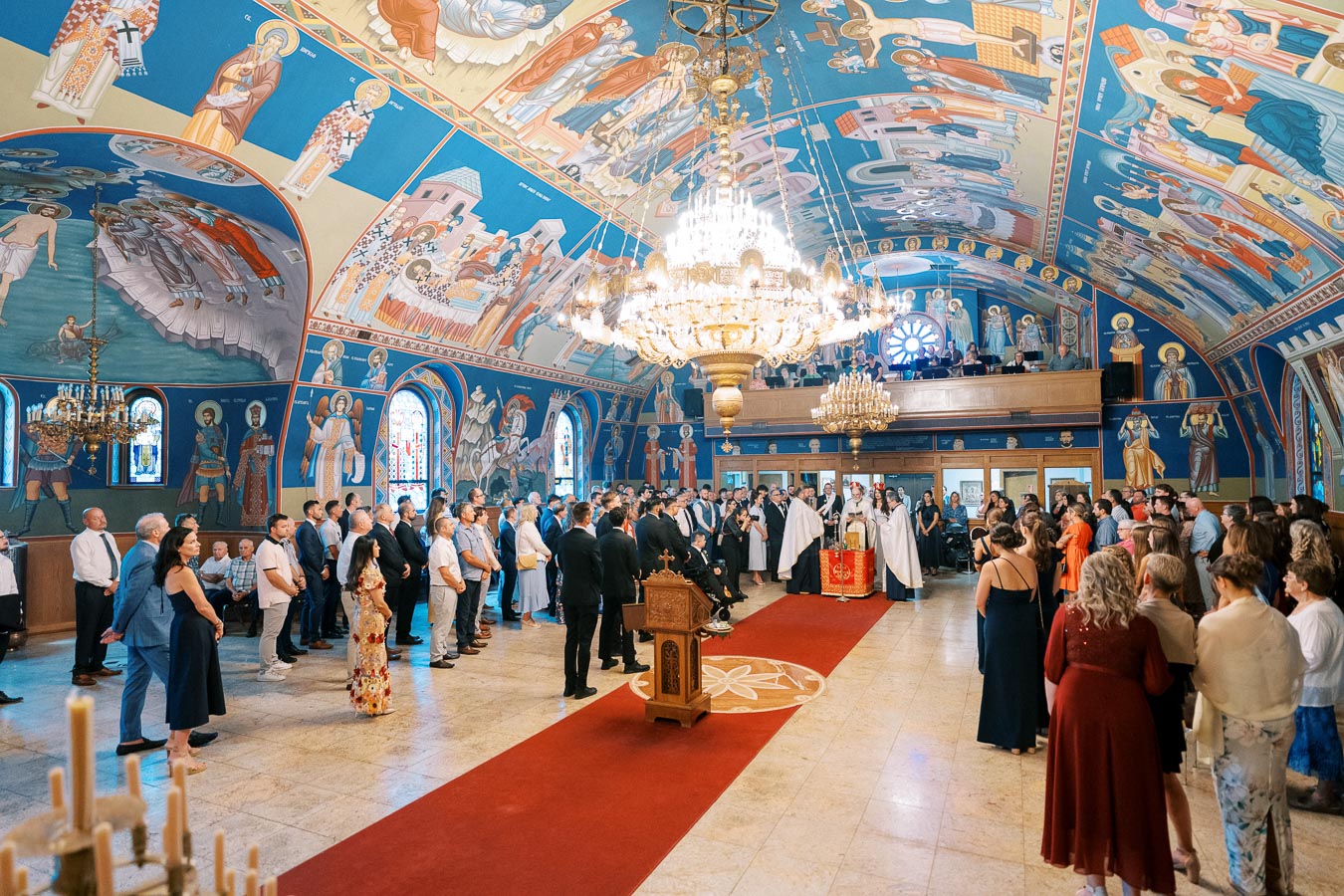 Church ceremony in a beautifully decorated hall with vibrant religious frescoes and ornate chandeliers, attended by a large gathering of people standing around a central altar on a red carpet.