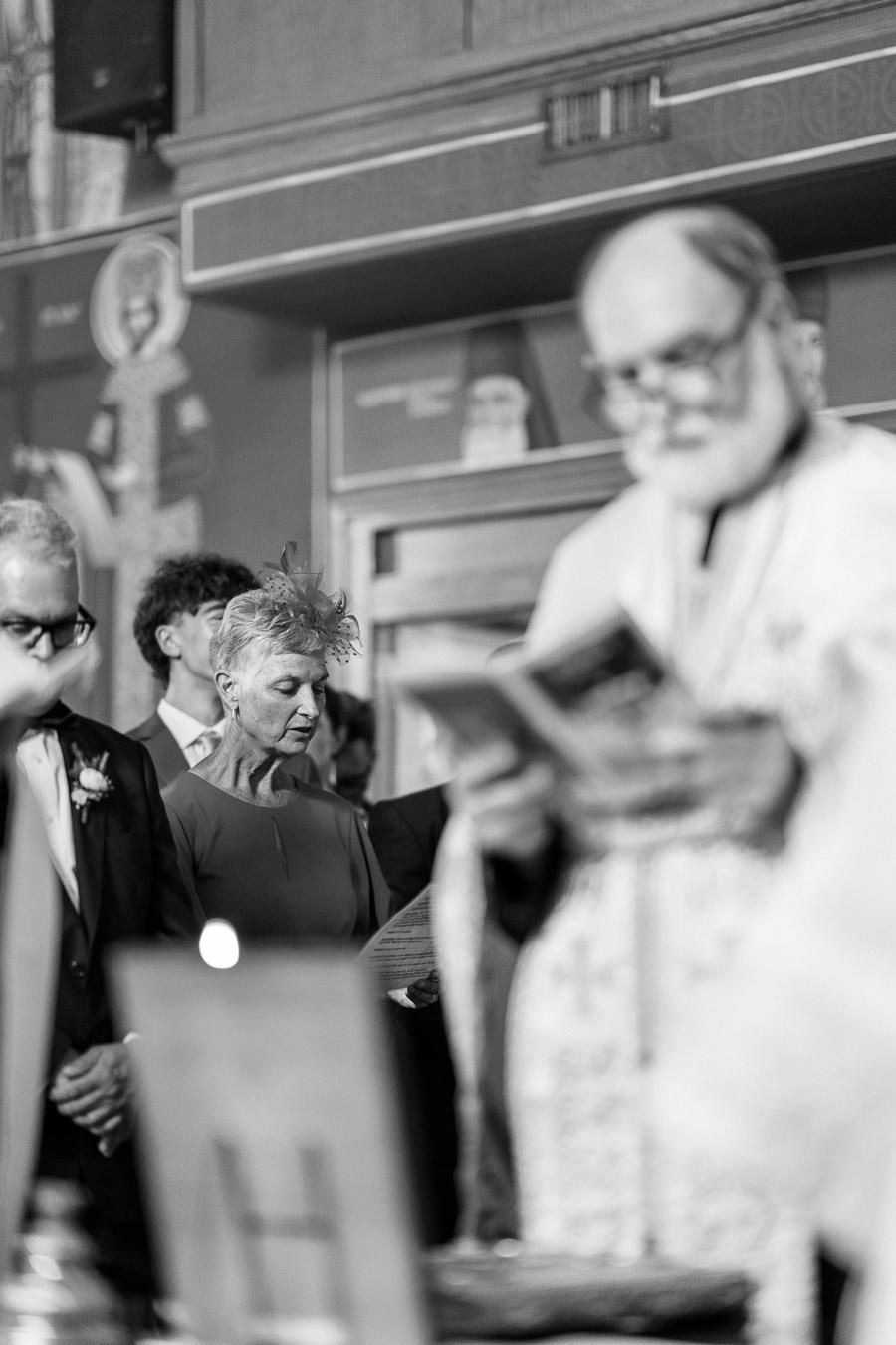 A black and white image of a solemn ceremony inside a church, with a priest holding a book in the foreground and several attendees in formal attire observing in the background.