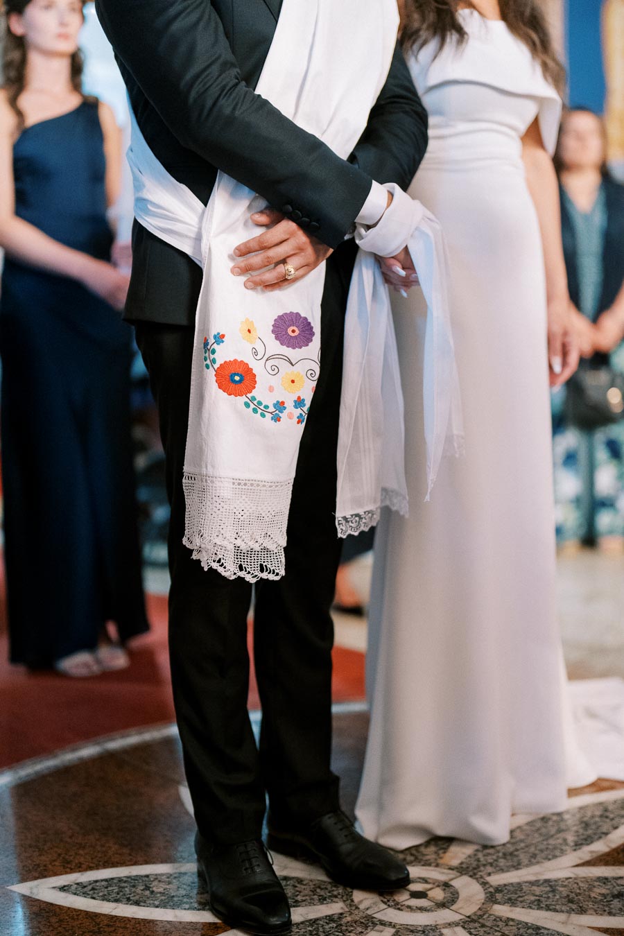 A couple stands during a wedding ceremony, with the groom wearing a black suit and a white scarf embroidered with colorful flowers and lace, while the bride wears a white gown.