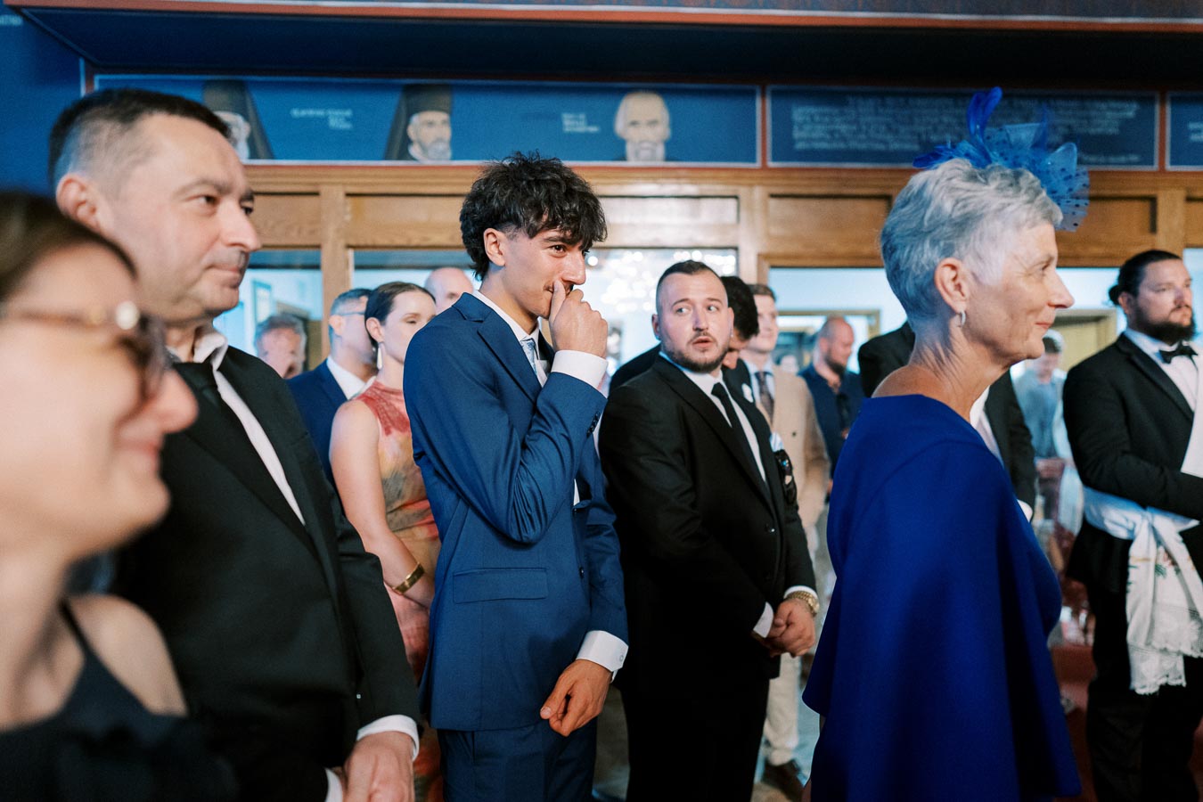 A group of elegantly dressed people at a formal event, with a man in a blue suit appearing thoughtful.