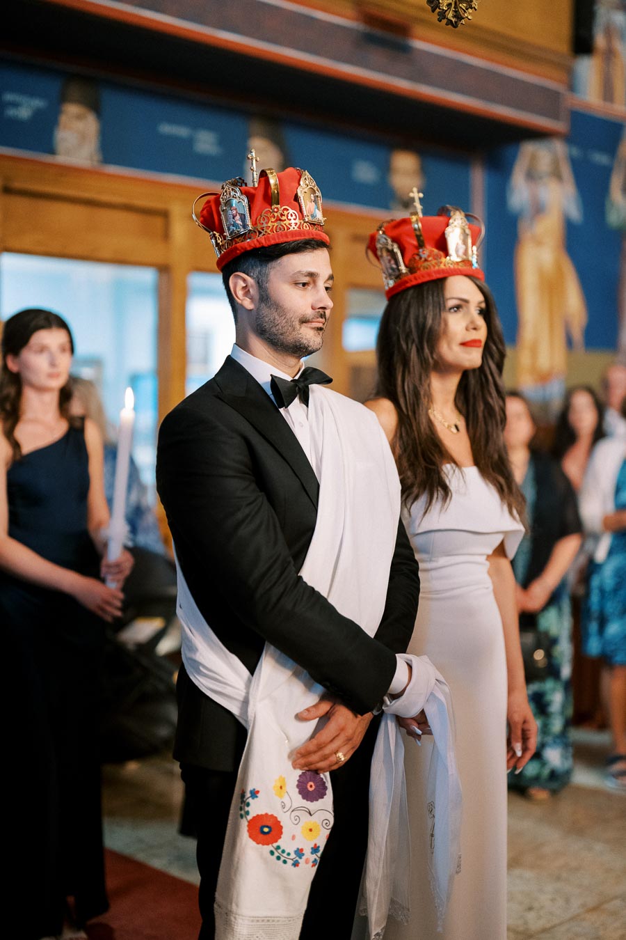 Couple in traditional wedding attire wearing ornate crowns during a religious wedding ceremony inside a decorated church.