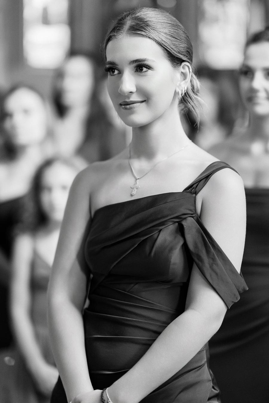 A young woman in a formal off-shoulder dress, standing elegantly at an event, with other attendees in the softly focused background.