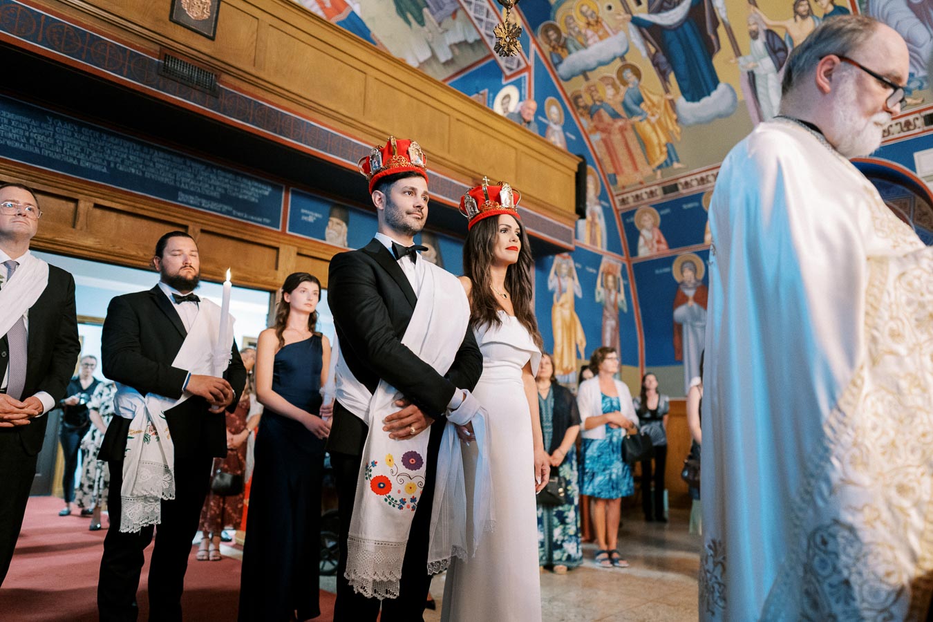 A bride and groom wearing red crowns stand together during a traditional wedding ceremony in an ornately decorated church interior, surrounded by formally dressed guests and a priest in white robes.