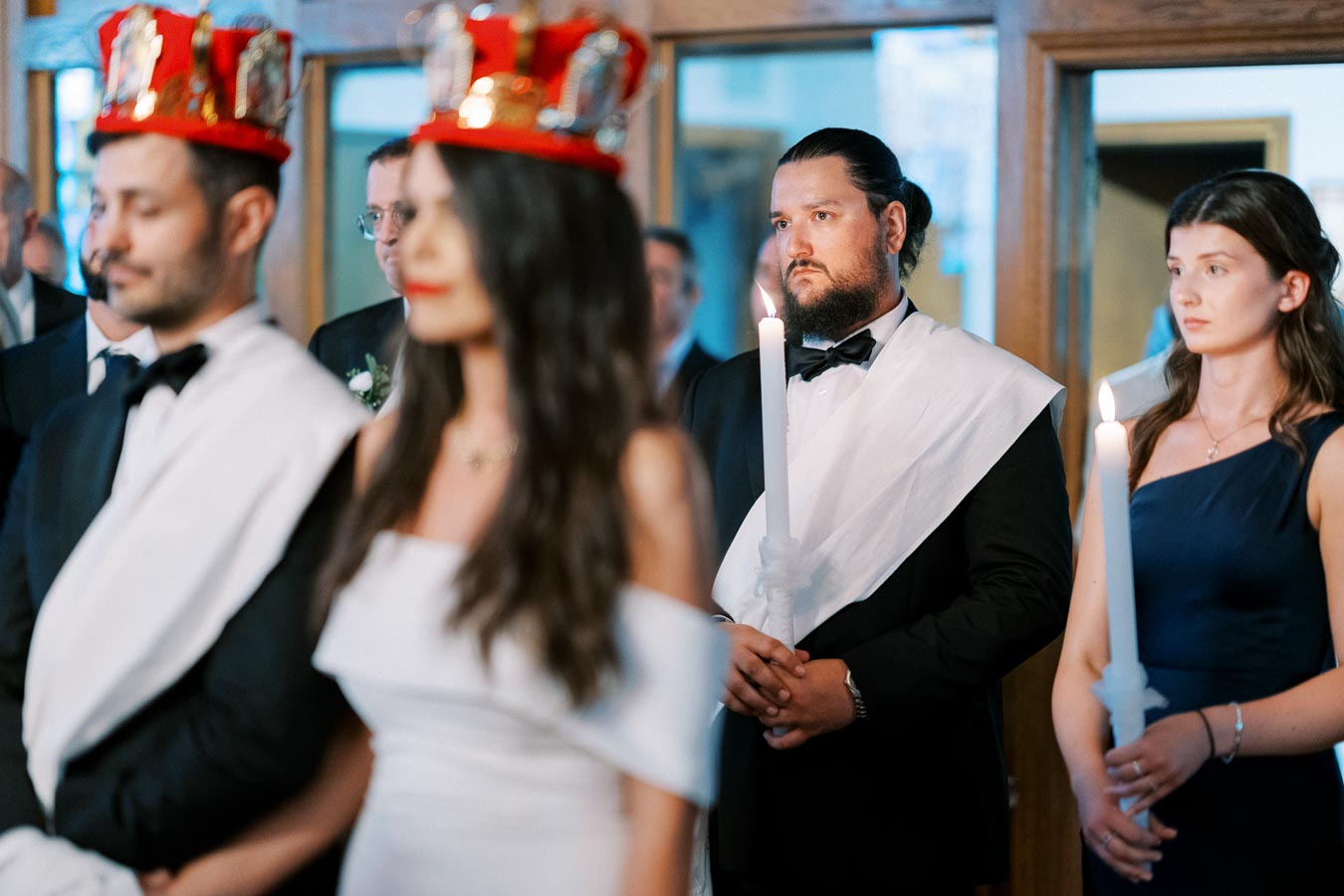A group of elegantly dressed individuals participating in a traditional ceremony, featuring men with red crowns and sashes, and women holding white candles, in a well-lit indoor setting.