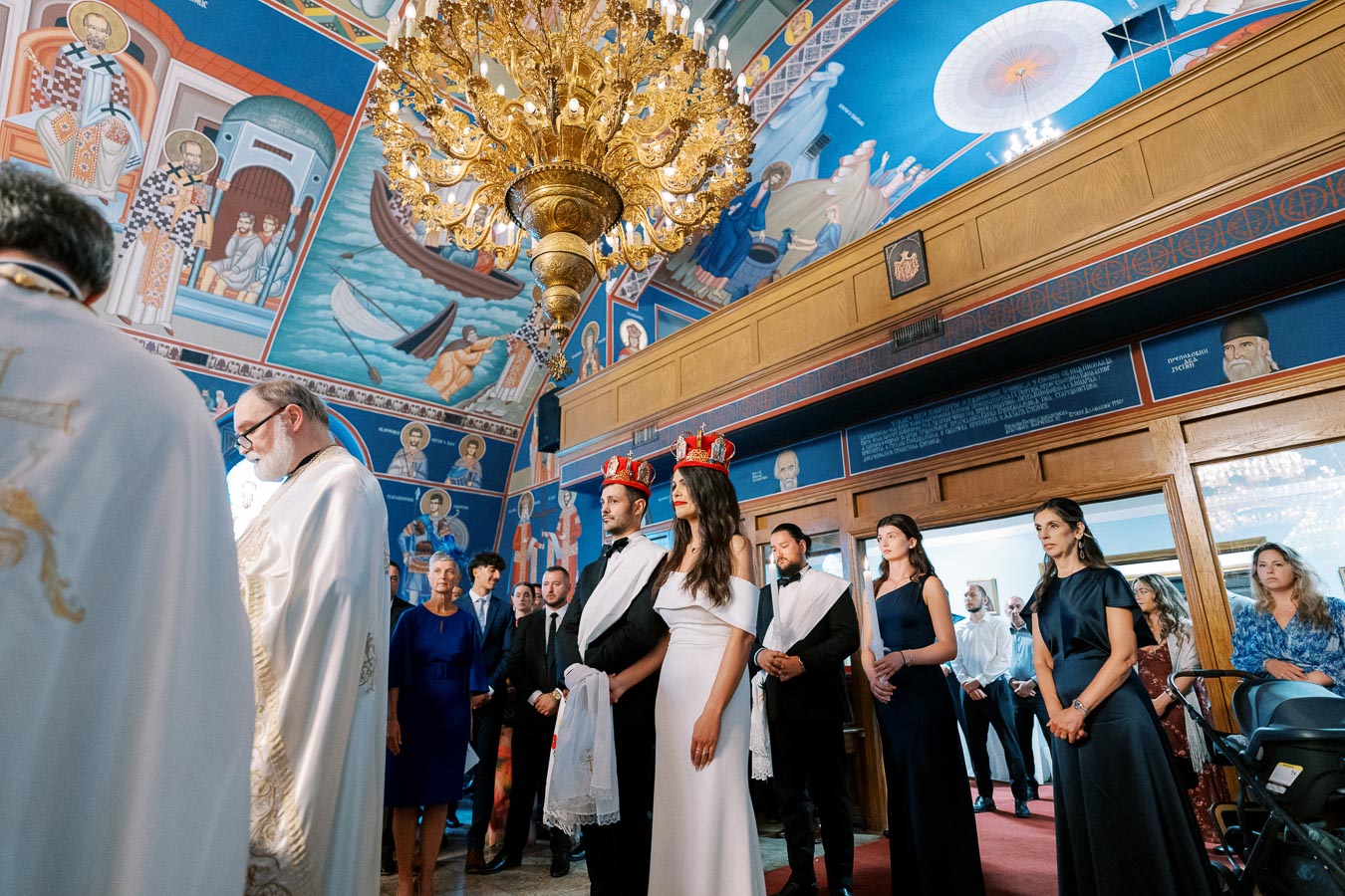 A traditional Eastern Orthodox wedding ceremony inside a beautifully decorated church, featuring a couple wearing red crowns. The ornate interior includes vibrant iconography and a grand chandelier, with guests gathered around witnessing the ceremony.