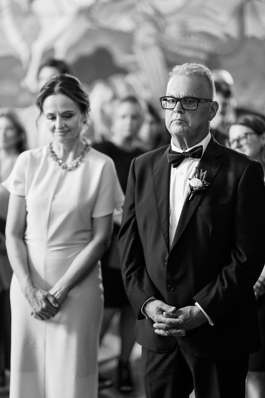 Black and white photo of a formally dressed older couple standing at an indoor event, with the man wearing a suit and bow tie, and the woman in a dress and necklace, surrounded by blurred guests in the background.