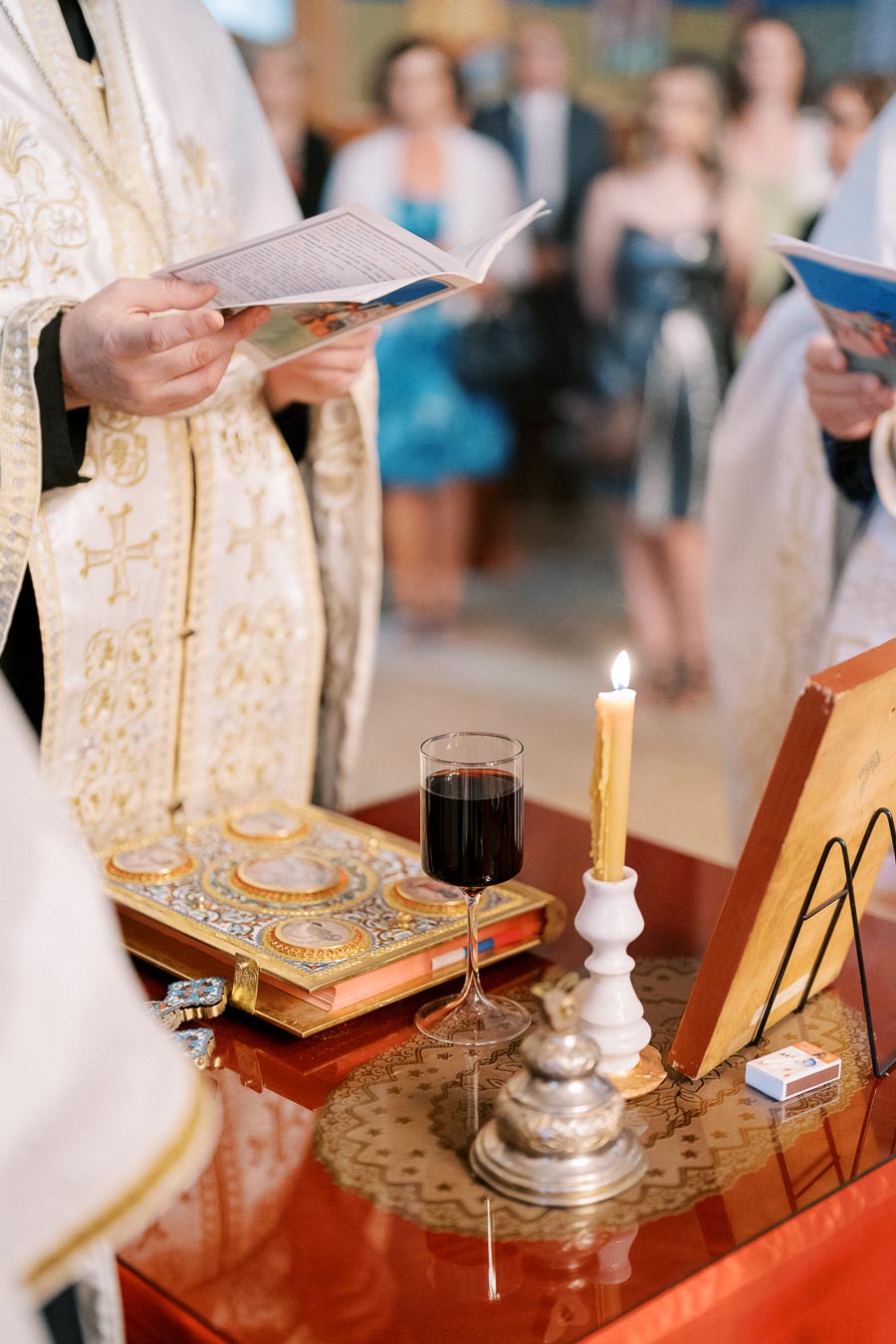 Certainly! Here is an SEO-friendly alt text for the image:Religious ceremony setup featuring a priest holding a book, an ornate religious text, a glass of wine, and a lit candle on a red table. Ceremony attendees blurred in the background, enhancing the solemn atmosphere.