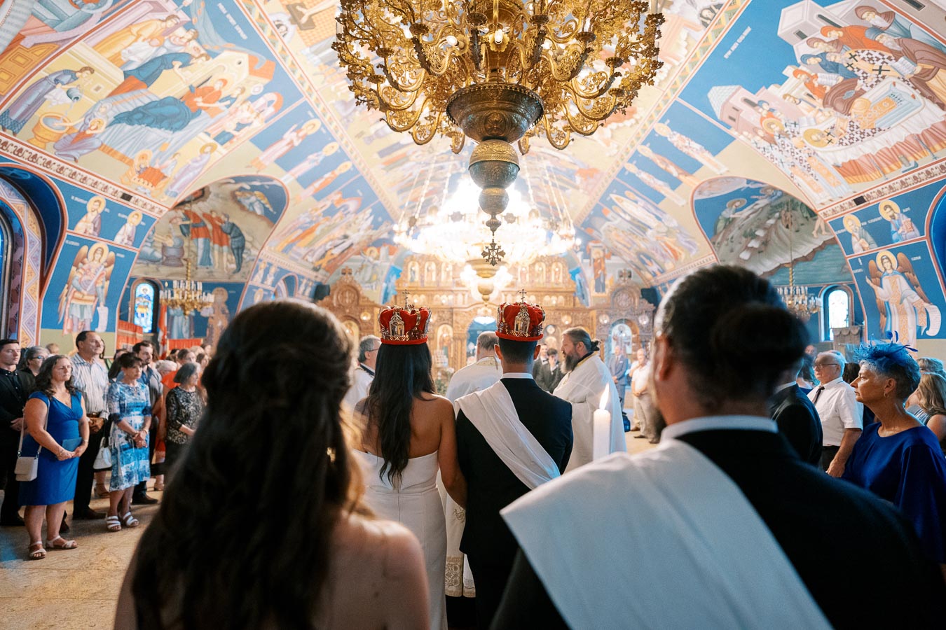 Ceremony inside an ornately decorated Orthodox church, featuring a bride and groom with crowns, attended by guests.
