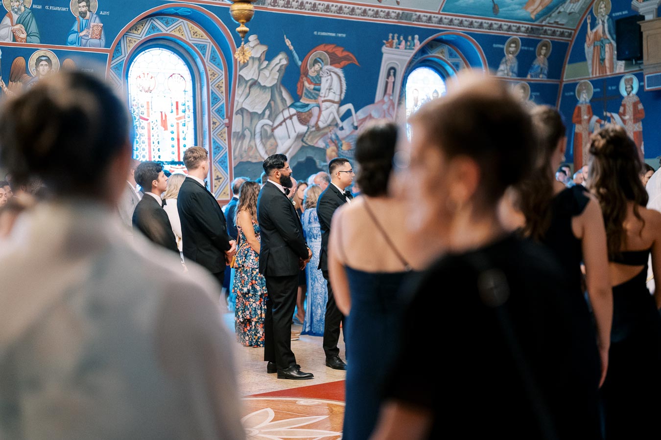 Guests standing inside a beautifully decorated Orthodox church, with vibrant wall murals and stained glass windows, during a formal event.