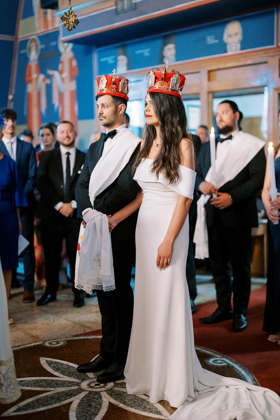 Couple wearing crowns during traditional church wedding ceremony, surrounded by guests in formal attire, with intricate religious decorations in the background.