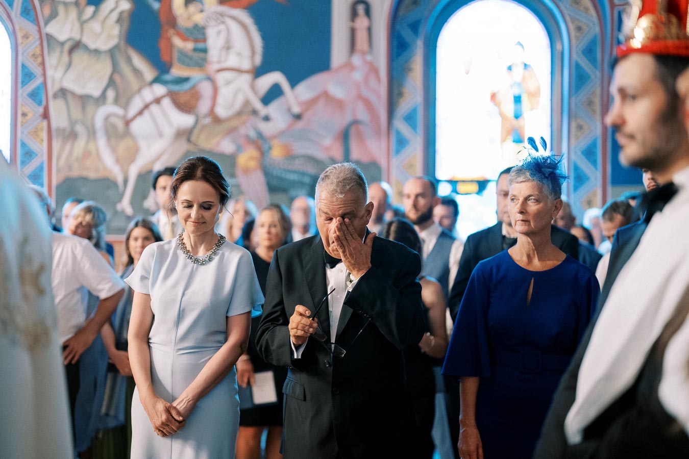 A group of people in formal attire attending an indoor ceremony, with an emotional elderly man wiping his eyes and vibrant mural artwork in the background.