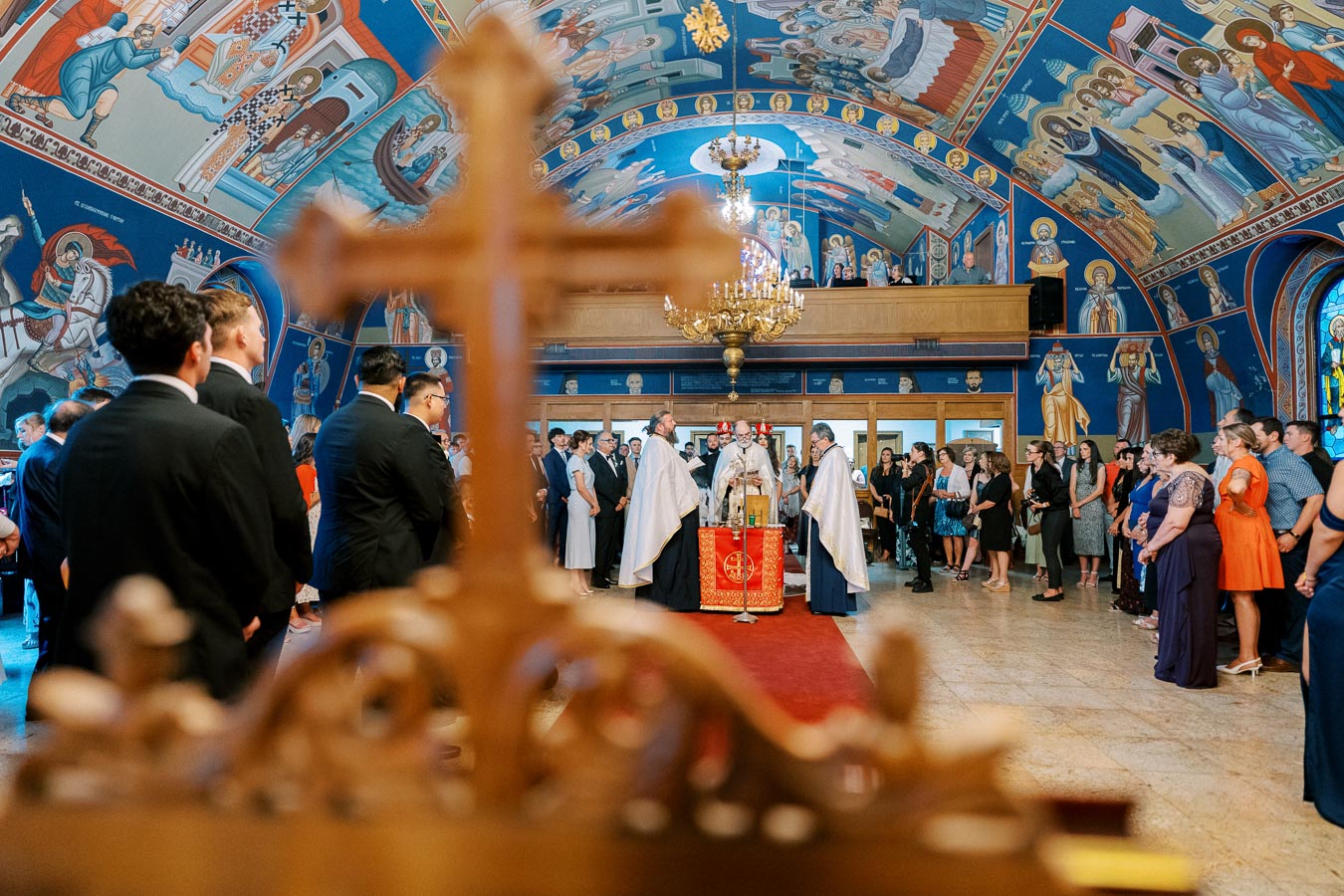 Orthodox church ceremony with priests in white vestments and attendees, surrounded by vibrant religious murals and a prominent chandelier.