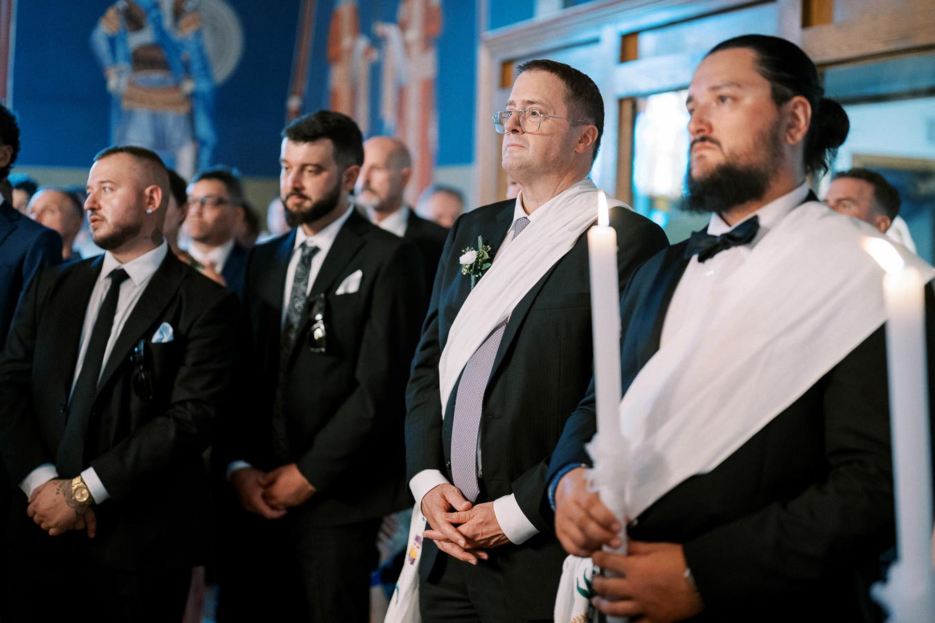 A group of men dressed in formal attire with white sashes, holding lit candles in a solemn ceremony inside a decorated venue.