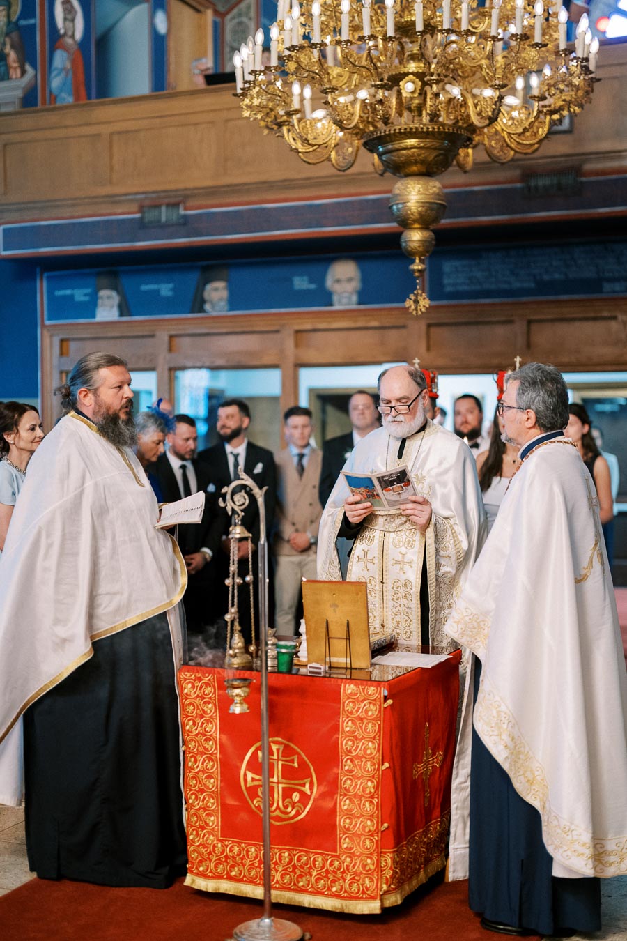 Orthodox priest conducting a religious ceremony inside a church, with ornate gold chandelier and religious artifacts on a red-covered table, surrounded by attendees in formal attire.