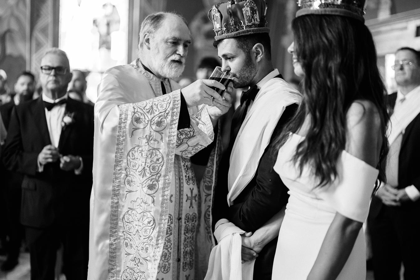 A black and white image of a traditional religious wedding ceremony, featuring a priest with ornate robes performing a ritual. The groom, wearing a crown and suit, is drinking from a ceremonial cup held by the priest. The bride stands beside him, wearing an off-shoulder gown and a matching crown. Guests are visible in the background.