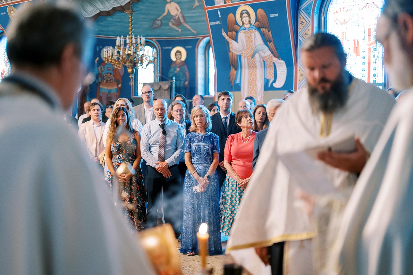 A congregation attentively participates in an Orthodox church service, with vibrant iconography displayed on the walls.