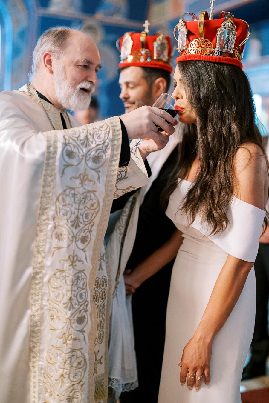 Orthodox wedding ceremony with priest offering wine to bride and groom wearing red crowns.