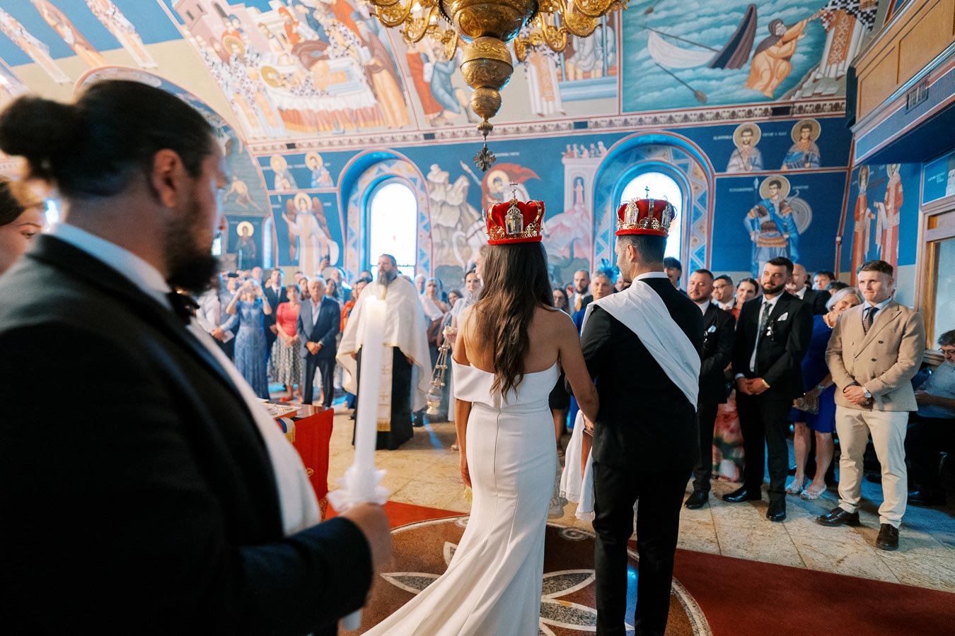 A bride and groom wearing crowns during a traditional wedding ceremony inside an ornately decorated church, surrounded by guests and vibrant murals.