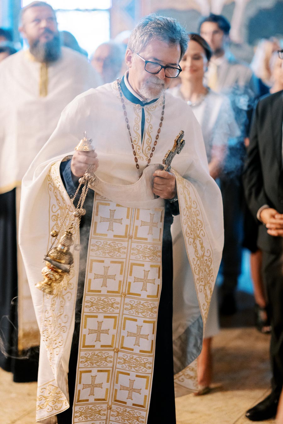 Priest in ceremonial attire holding a cross and censer during an Orthodox church service, with blurred congregation in the background.