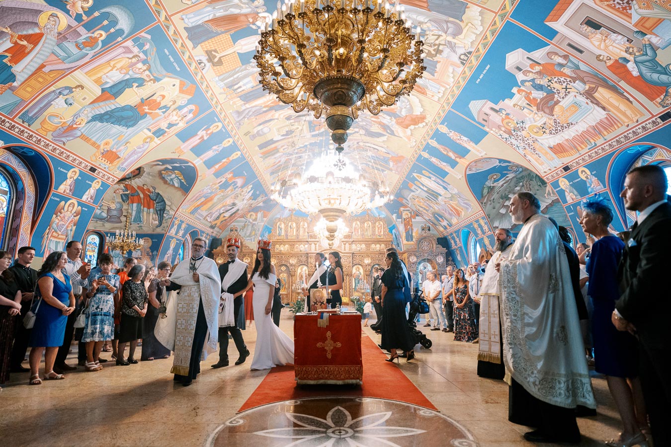 A vibrant wedding ceremony inside an ornately decorated church, featuring colorful frescoes and a grand chandelier. A priest leads a couple dressed in traditional attire down the aisle, surrounded by family and guests.