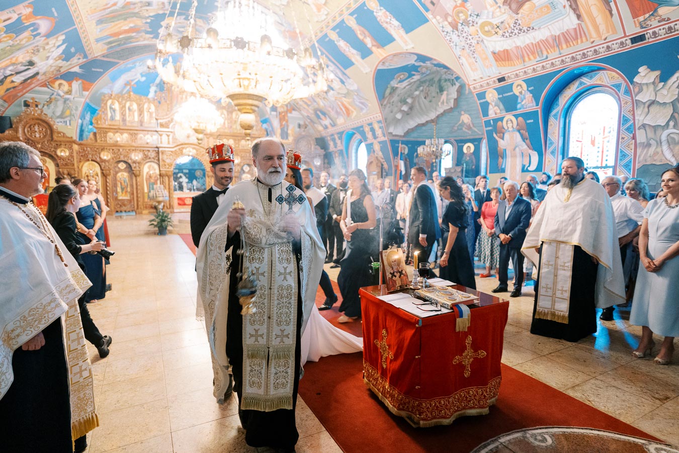 Priest conducting an Orthodox wedding ceremony inside an ornate church with colorful frescoes and a large chandelier, as guests and a couple wearing crowns look on.