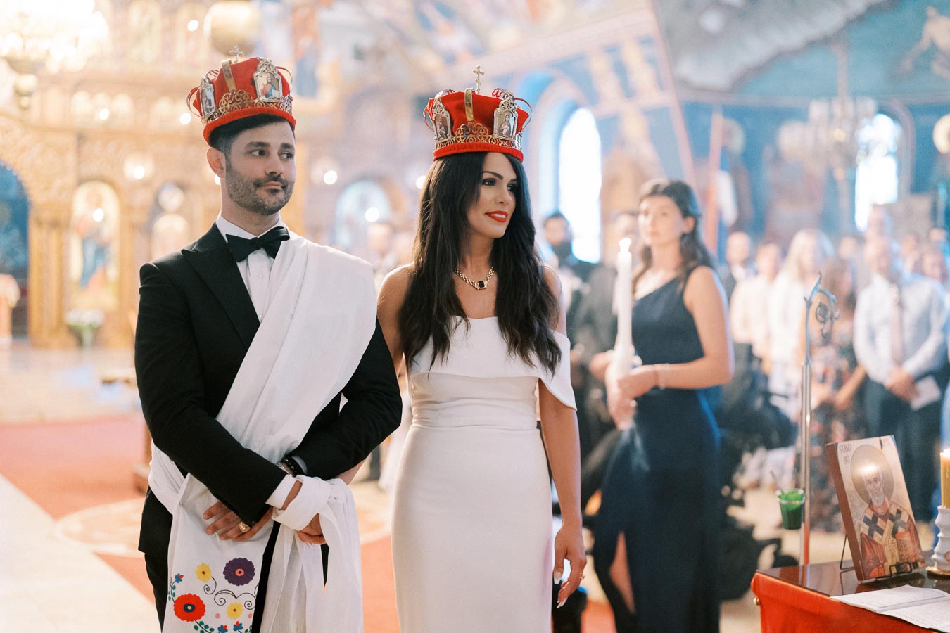 A bride and groom in formal attire and crowns stand in a beautifully decorated church during their wedding ceremony, with guests in the background.