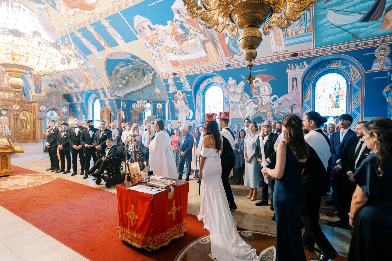 A traditional church wedding ceremony featuring a couple standing before a priest, surrounded by guests inside an ornately decorated church with colorful murals and stained glass windows. The bride wears a white dress and the groom is in formal attire.