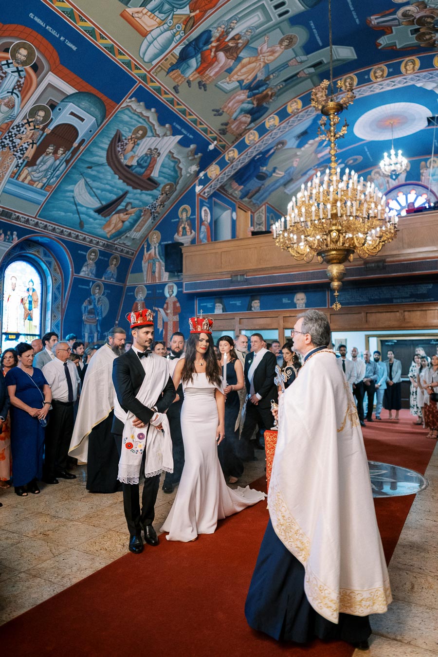 A couple in formal attire stands at the altar during a traditional Greek Orthodox wedding ceremony, surrounded by family and friends in a beautifully decorated church with vibrant religious murals and a large chandelier.