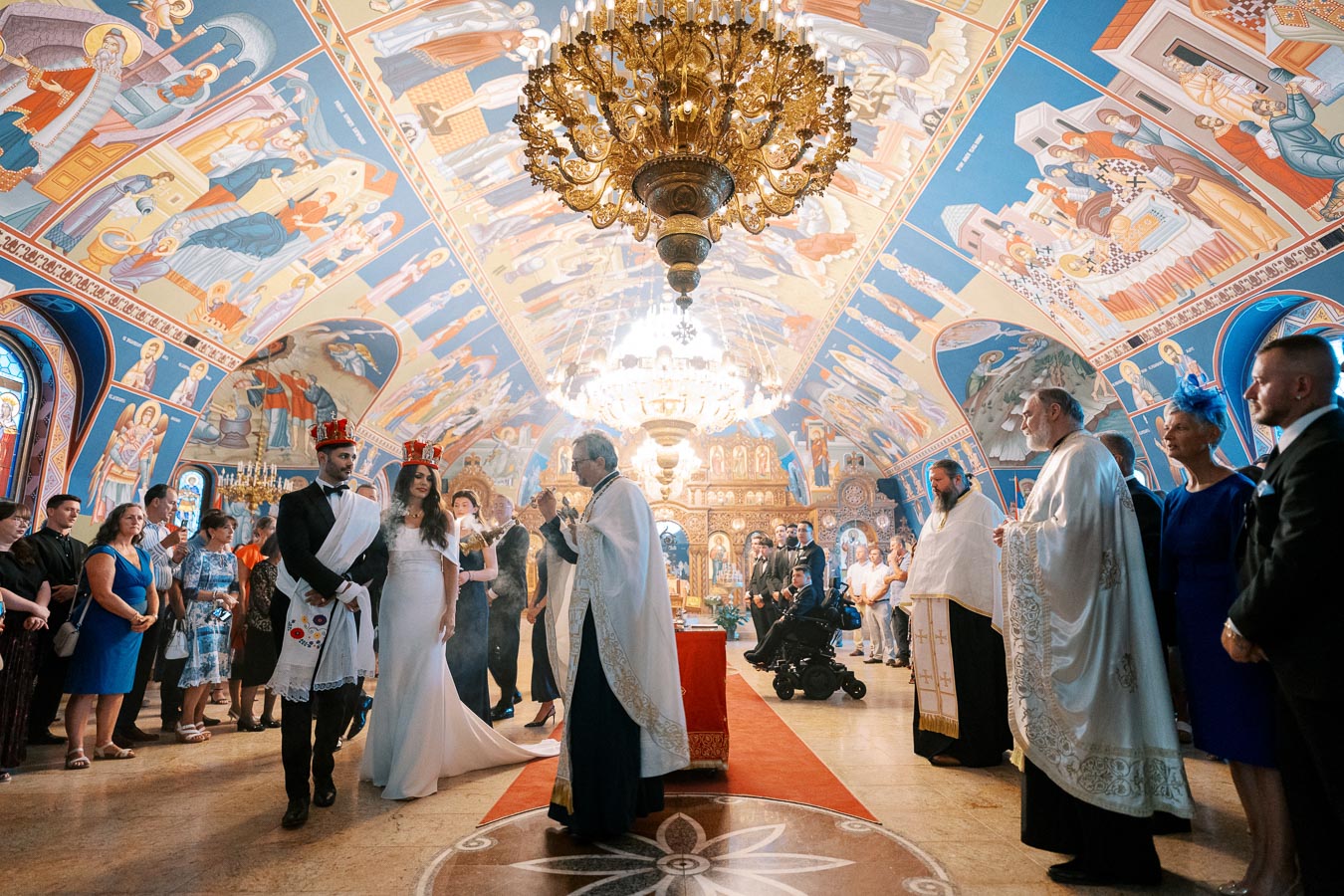 Orthodox wedding ceremony in beautifully decorated church with intricate frescoes and golden chandeliers.